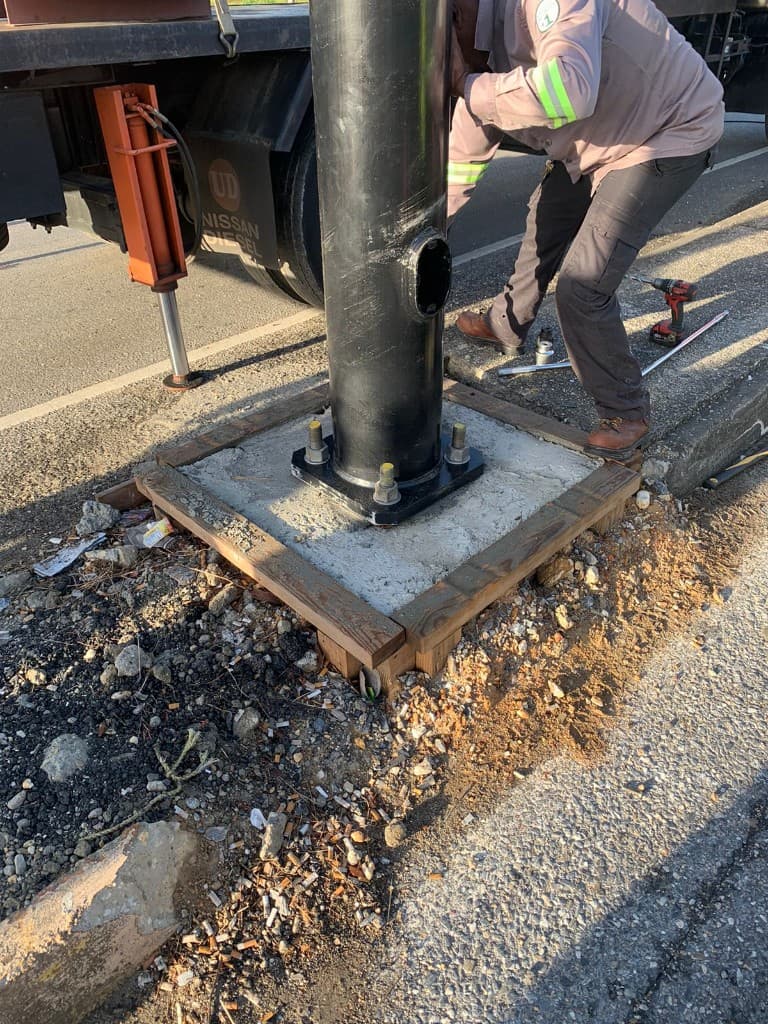 Technician working at the base of a traffic signal mast anchored to a concrete foundation beside a service vehicle